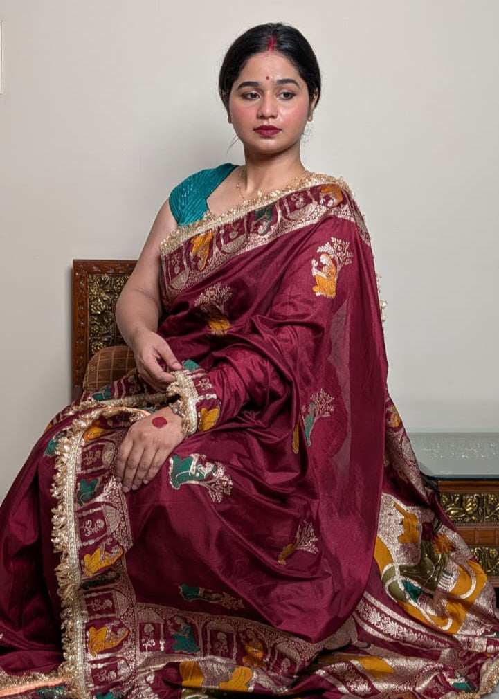 Woman in a traditional maroon saree with gold embroidery sitting on a chair.